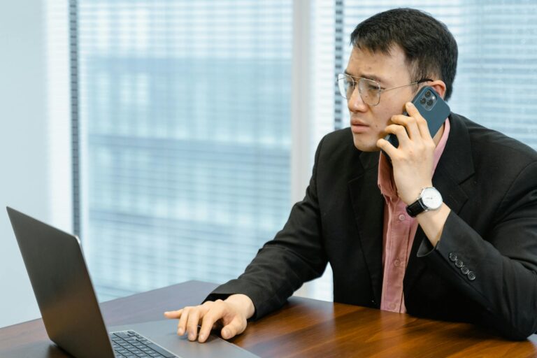 Asian businessman wearing blazer, working with laptop and smartphone in modern office.